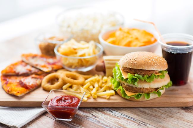 Na dřevěném podnosu je naservírováno jídlo, které způsobuje reflux a pálení žáhy, jako je hamburger, pizza, chipsy, kečup, smažená jídla a coca cola.